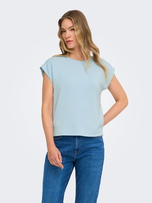 Kvinde med langt blond hår iført en lyseblå kortærmet top og blå jeans, stående med den ene hånd på hoften mod en ensfarvet baggrund.
