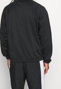 Persoon in een zwart jack met lange mouwen en zwarte broek met witte zijstreep, gezien vanaf de achterkant tegen een effen achtergrond.