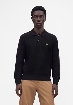 Giovane uomo che indossa una polo nera a maniche lunghe con un piccolo logo verde sul petto e pantaloni beige, in piedi davanti a uno sfondo semplice.