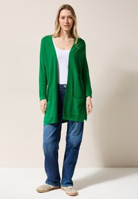 Grön kofta med långa ärmar, öppen front och två framfickor, parat med blå denimjeans och ljusa skor.