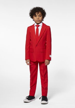 Traje rojo con chaqueta ajustada y corbata a juego, camisa blanca debajo y un solo bolsillo. Combinado con zapatillas negras para un contraste.