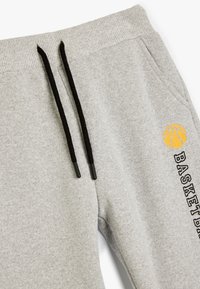 Pantalones de chándal grises con cordones negros, que presentan un gráfico de baloncesto y el texto "BASKETBALL" en negro en la pierna izquierda. Textura suave.
