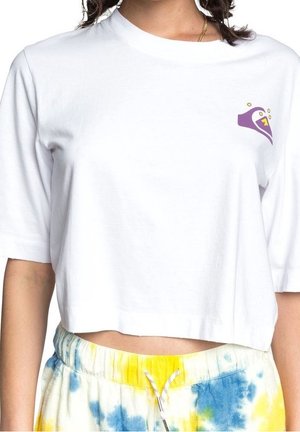 Frau trägt ein weißes, kurzes T-Shirt mit violettem Motiv auf der Brust und gelb-blauen Tie-Dye-Shorts mit Kordelzug, sichtbare goldene Halskette.