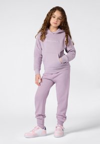 Junges Mädchen mit lockigem Haar, das einen lila Kapuzenpullover und passende Sweatpants trägt, steht mit einer Hand in der Tasche und pinken Turnschuhen.