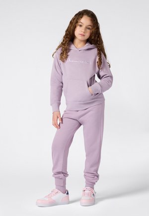Meisje met krullend haar dat een lavendelkleurige hoodie en bijpassende sweatpants draagt, staande met één hand in de zak en roze sneakers.