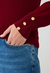 Maglione a maglia bordeaux con polsini a coste, caratterizzato da tre bottoni dorati. Abbinato a jeans in denim blu, che mostrano un dettaglio della tasca.