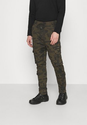 Personne portant un pantalon cargo camouflage vert foncé et noir, des chaussures montantes noires et un haut à manches longues noir, sur un fond uni.