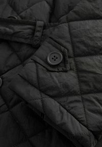 Gros plan sur la manche d'une veste matelassée noire avec un motif losange cousu et un seul bouton noir sur une petite patte.