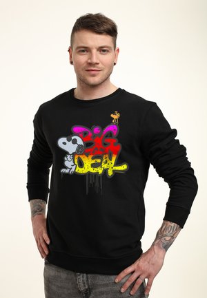 Jeune homme aux cheveux courts et bruns, debout les mains sur les hanches, portant un sweat-shirt noir avec un design graffiti coloré de Snoopy et Woodstock où il est écrit Big Deal.
