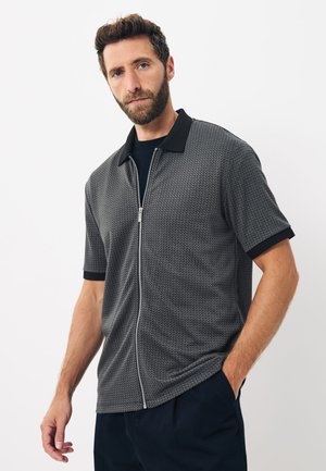 Hombre con barba que lleva una camisa corta de manga con cremallera de patrón gris, con cuello y puños negros, de pie ante un fondo blanco liso.