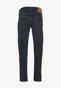 Jean en denim bleu foncé vu de dos avec deux poches arrière cousues et un patch de marque en cuir marron sur la ceinture.