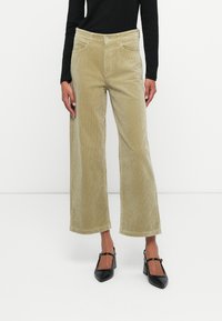 Wijd-gelopen, corduroy broek in licht olijfgroen met een textuurafwerking. Voorzien van twee voorzakken en een knoopsluiting.