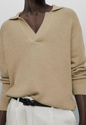 Osoba o ciemnej karnacji ubrana w beżowy prążkowany sweter z dekoltem w serek i białe spodnie z czarnym paskiem, ramiona swobodnie opuszczone wzdłuż ciała.