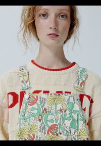 Młoda kobieta o kręconych rudych włosach, ubrana w kremowy sweter z czerwonym napisem oraz ogrodniczki w kwiatowy wzór z zielonymi, żółtymi i czerwonymi detalami.