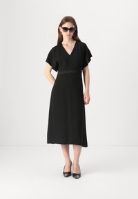 Črna obleka s kratkimi krili, v-izrezom in detajlom pasu. Ujemajoče se z črnimi visokimi petami in oversized sončnimi očali.