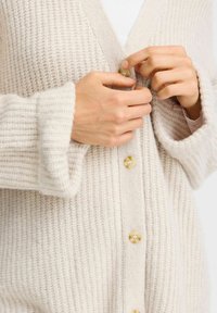 Cardigan lavorato a maglia di un beige chiaro con texture a costine, dotato di scollo a V, maniche lunghe e grandi bottoni crema lungo la parte anteriore.