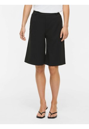 Kvinde iført sorte knælange vidde shorts og sorte hælede sandaler, stående mod en ensfarvet lys baggrund.
