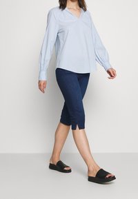 Vaaleansininen, pitkähihainen paita V-pääntiellä, yhdistettynä tummiin farkkulegginseihin, joissa on sivurei'ät, ja mustiin liukusandaaleihin.