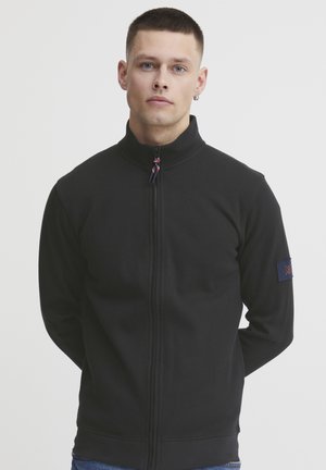 Chaqueta negra con cremallera y cuello alto, de tejido texturizado, y un parche azul con un emblema en la mangas izquierda.