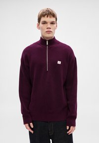 Μπορντό πλεκτό πουλόβερ με ψηλό γιακά, μισή φερμουάρ και μικρό λογότυπο στην αριστερή πλευρά του στήθους. Διαθέτει μακριά μανίκια και ριμπ υφή.