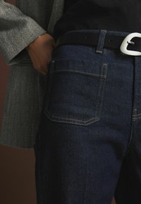Jeans in denim blu scuro con un design fitted, caratterizzati da cuciture beige e una tasca posteriore. È visibile anche una cintura nera con una fibbia argentata.