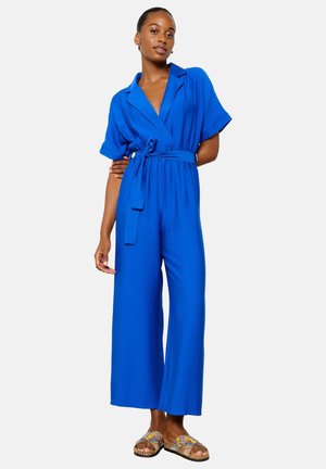 Monos azul con mangas cortas, piernas anchas y un cinturón en la cintura. Tela ligera con una textura suave. Combinado con sandalias estampadas.