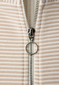 Světle béžová a bílá pruhovaná tkanina se stříbrným zipem, vybavená kulatým jezdcem a texturovaným pletením. Pohled zblízka.
