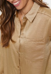 Hellgoldenes Hemd aus glänzendem Stoff mit Brusttasche und spitzem Kragen, versehen mit braunen Knöpfen. Glatte Textur.