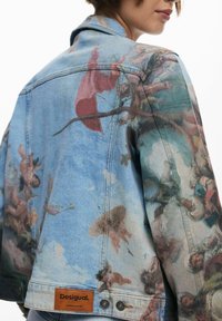 Veste en denim avec un design coloré et pictural représentant des nuages et des silhouettes. Base bleue, étiquette en cuir marron et accents en métal.