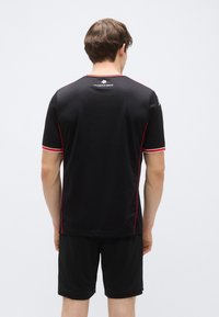 T-shirt de sport noir avec des accents rouges et une texture côtelée, manches courtes et col rond. Le mannequin porte un short noir.