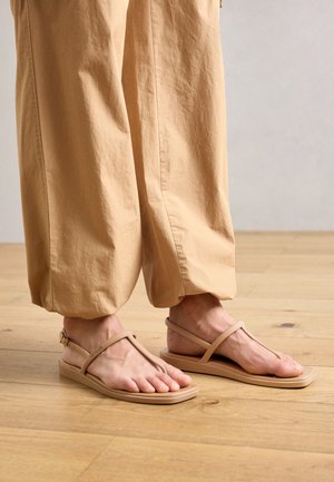 Beige Zehentrenner mit flachen Sohlen, minimalistisches Design und schmalen Riemen, kombiniert mit lockeren, hellen Hosen auf einem Holzboden.