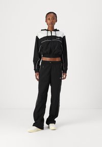 Chándal en blanco y negro con chaqueta con capucha corta y pantalones sueltos. Presenta puños y cinturilla elásticos, con acentos laterales blancos y cremalleras.