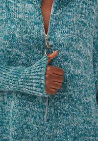 Ręczne zaciąganie suwaka w postaci srebrnego pierścienia na turkusowym i białym swetrze z długimi rękawami, które zakrywają nadgarstek i część dłoni.