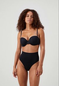 Woman with curly hair wearing a black strapless bra with removable straps and high-waisted black underwear, standing against a plain background.