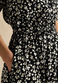 Schwarzes Kleid mit weißem Blumenmuster, elastischem Taillenbund, kurzen Ärmeln und Seitentaschen. Weicher Stoff mit lockerer Passform.