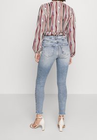 Jeans de denim azul de cintura alta con una textura desgastada, dobladillo deshilachado y bolsillos traseros. Combinados con una blusa estampada y tacones.