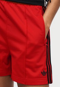 Κόκκινα αθλητικά σορτς από λείο ύφασμα, με μαύρες λωρίδες στα πλάγια και κεντημένο μαύρο λογότυπο στο κάτω αριστερό μέρος. Σχεδιασμένα για άνεση.