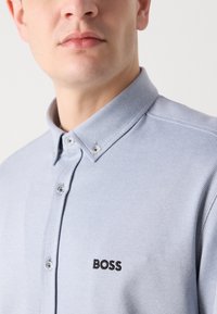 Šviesiai mėlynas tekstūruotas marškinis su mygtukais ant apykaklės, su juodu siuvinėtu "BOSS" logotipu ant krūtinės.