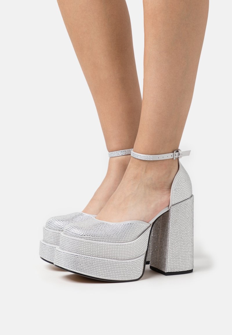 Escarpins à plateforme argentée avec brides à la cheville, ornés de petits strass, portés sur des pieds nus sur fond blanc.