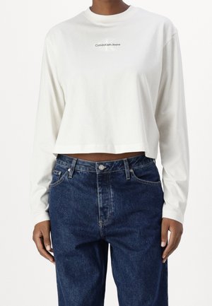 Osoba má na sobě bílý krátký top s dlouhým rukávem s logem "Calvin Klein Jeans" a tmavě modré vysoké denimové džíny.