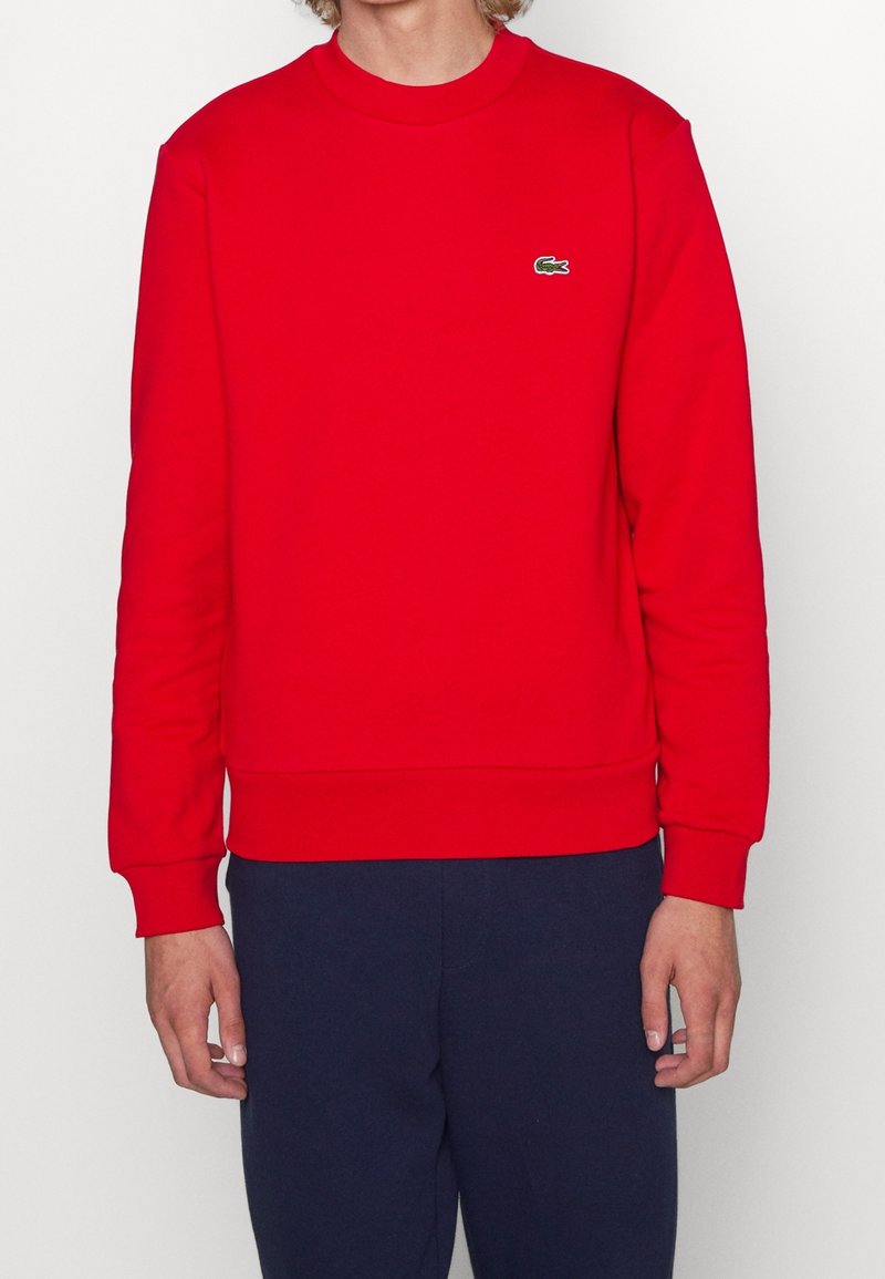 Personne portant un sweat-shirt rouge uni à manches longues avec un petit logo de crocodile vert sur la poitrine et un pantalon bleu foncé.