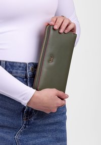 Olive green leather wallet with a gold logo, zip closure, and red accent. Held in hand, featuring a smooth texture and rectangular shape.