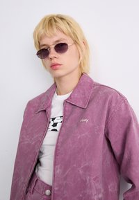 Rosa denimjakke med teksturert finish, frontglidelås og "obey"-logo. Brukt over en hvit skjorte med svart mønster, sammen med runde lilla solbriller.