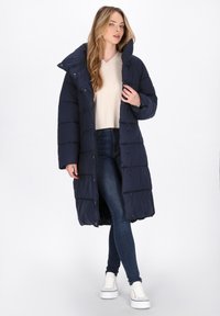 Cappotto trapuntato blu navy con colletto alto, chiusura frontale con bottoni e maniche lunghe, abbinato a un maglione leggero e jeans.