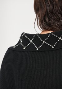 Fekete kötött pulóver bordázott textúrával; nyakán átlátszó cirkónia gyémántmintával díszített gallér található.