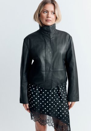 Kvinde med kort blond hår iført en sort læderjakke med høj krave og et sort polkaprikket skørt med blondekant.