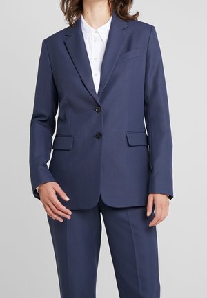Femme portant une veste de costume bleu marine à deux boutons et un pantalon assorti, par-dessus une chemise blanche à col, debout devant un fond gris.