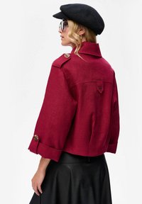 Rode linnen cropped jasje met knoopdetails op de mouwen, rug yoke en een rond kraag. Gekozen met een zwarte platte pet en een zwarte rok.