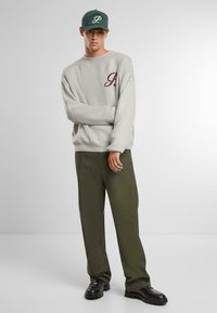Suéter de punto gris con un logo marrón, gorra verde, pantalones verde olivo y zapatos negros gruesos. El conjunto presenta líneas sencillas y casuales.