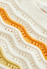 Tissu tricoté blanc avec des vagues texturées en rayures jaunes et orange, montrant des motifs de points détaillés et une étiquette de vêtement partielle.